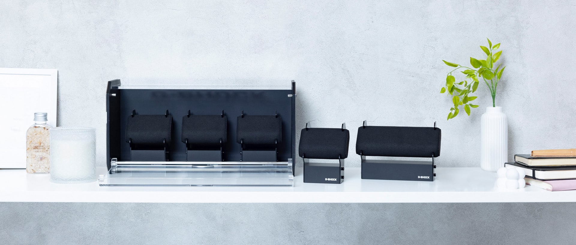 Três mostradores de relógios diferentes e a caixa de relógio da G-SHOCK numa mesa ao lado de uma jarra e de cadernos.