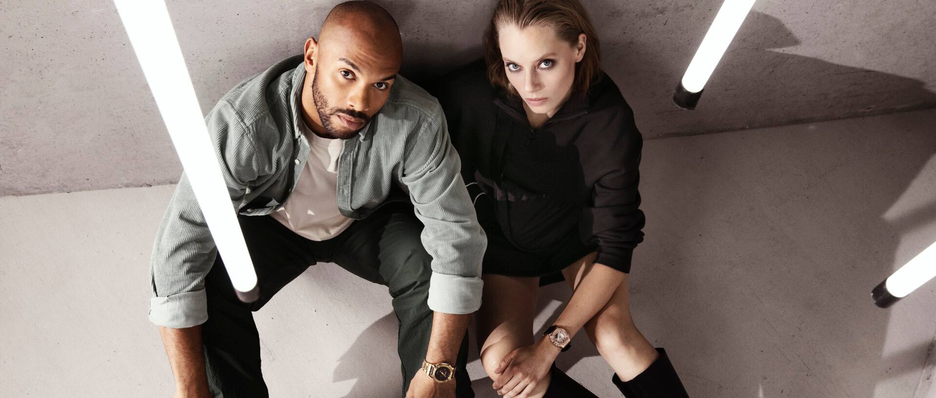 Duas pessoas estão sentadas junto a um muro de betão, com bastões luminosos pendurados ao lado delas. Ambas têm relógios de pulso Casio G-Shock da série GM-S2100.