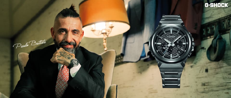 Paulo Battista, vestindo um terno preto, está sentado em uma poltrona de leitura ao lado de um abajur e usa o G-SHOCK G-STEEL GST-B1000. O relógio também está em destaque na imagem.