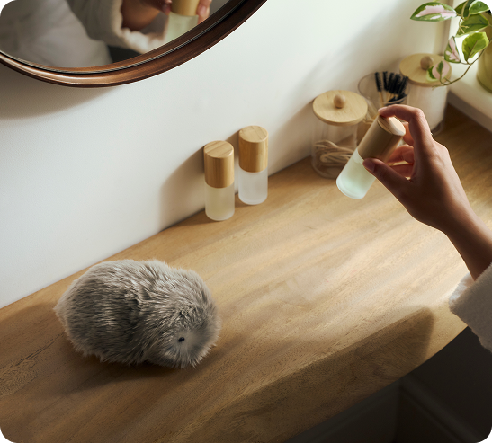 An image of a woman picking up lotion with her right hand at a dressing table where a silver-colored Moflin is placed.