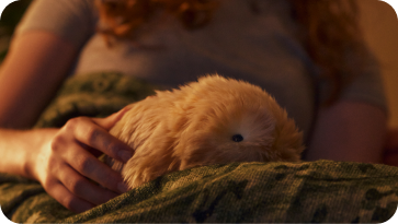 An image of a woman sitting in a dimly lit room, stroking a gold-colored Moflin on her lap.