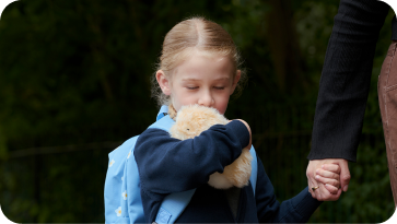 An image of a girl hugging a gold-colored Moflin with her right arm, pressing her face against it, while holding hands with her mother using her left hand.
