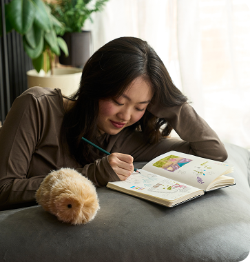 An image showing a person resting their head on their left hand with their elbow on the table, writing notes in a book with a pen in their right hand, while a gold-colored Moflin sits nearby.