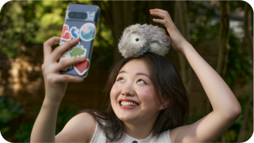 An image of a woman placing a silver-colored Moflin on her head with her left hand while taking a selfie with her smartphone in her right hand.