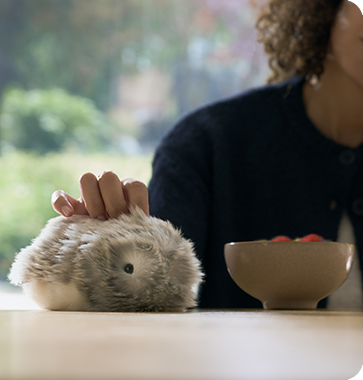 An image of a woman touching a silver-colored Moflin on a table with her right hand.