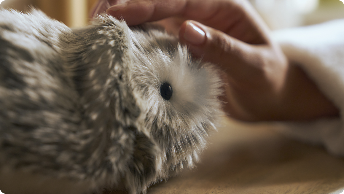 An image of a silver-colored Moflin being stroked on its face with a right hand.