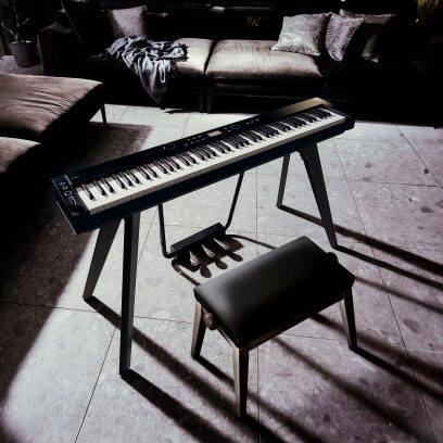 Aufnahme des CASIO Privia PX-S7000 Digitalpianos mit Sitzbank in Schwarz in einem dunkel gehaltenen Wohnraum mit Sofa und Steinboden, geprägt von starkem Lichteinfall mit ausgeprägten Schatten und hohem Kontrast.