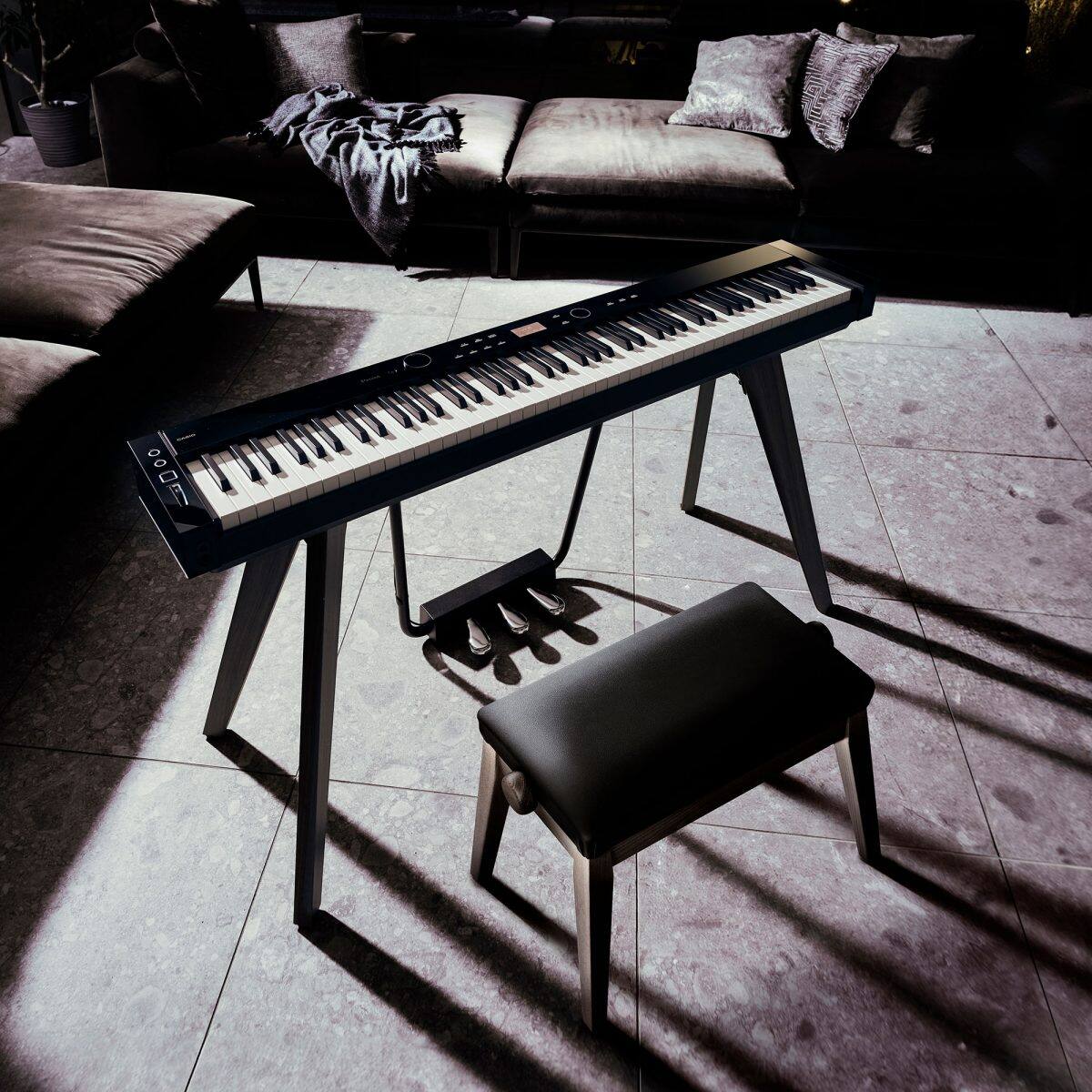 Aufnahme des CASIO Privia PX-S7000 Digitalpianos mit Sitzbank in Schwarz in einem dunkel gehaltenen Wohnraum mit Sofa und Steinboden, geprägt von starkem Lichteinfall mit ausgeprägten Schatten und hohem Kontrast.
