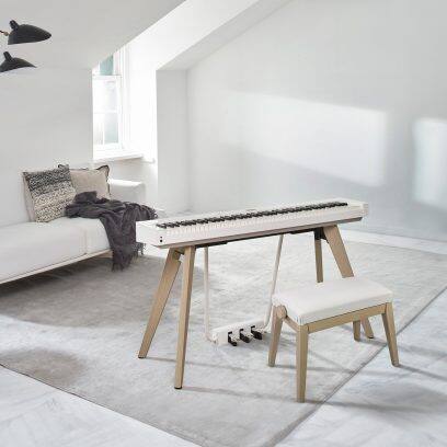 Aufnahme des CASIO Privia PX-S7000 Digitalpianos mit Sitzbank in Weiß in einem modern eingerichteten, hellen Wohnraum mit Sofa und Teppich, bei einfallendem Licht durch ein Fenster.