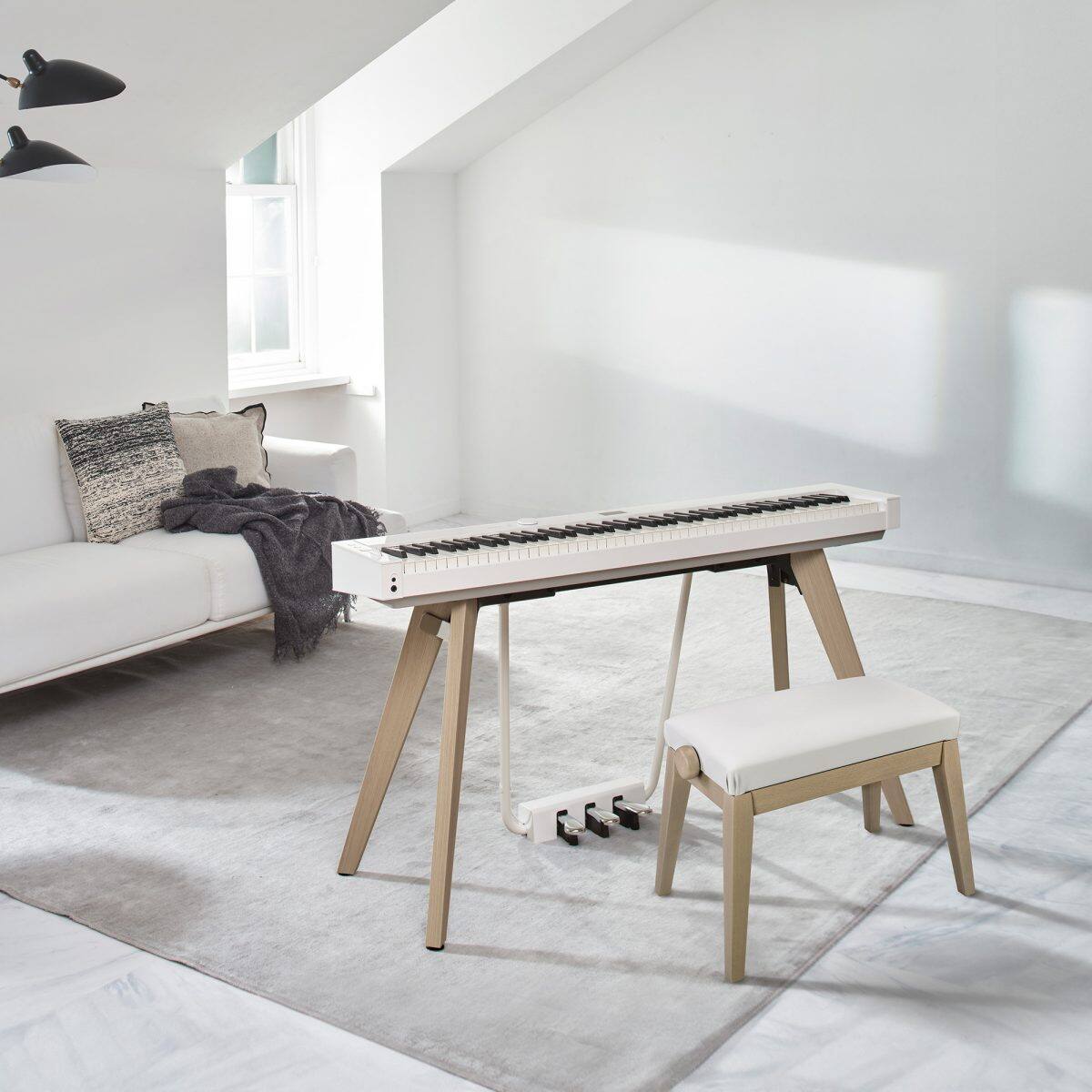 Aufnahme des CASIO Privia PX-S7000 Digitalpianos mit Sitzbank in Weiß in einem modern eingerichteten, hellen Wohnraum mit Sofa und Teppich, bei einfallendem Licht durch ein Fenster.