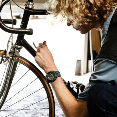 Ein Mann schraubt an einem Fahrrad, an seinem Handgelenk ist die dunkelgrüne G-SHOCK GA-010-2A Armbanduhr zu sehen.