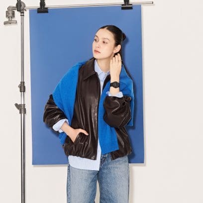 Eine Frau mit einer schwarzen Lederjacke und einem blauen Strickpullover trägt die GMA-P2100BA-1AER am Handgelenk und steht vor einer blauen Leinwand