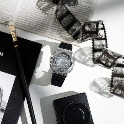 Die GM-S2110-1A Armbanduhr liegt auf einem Tisch neben einem Fotofilm, einer Kamera, einer Zeitung und einem Bleistift.