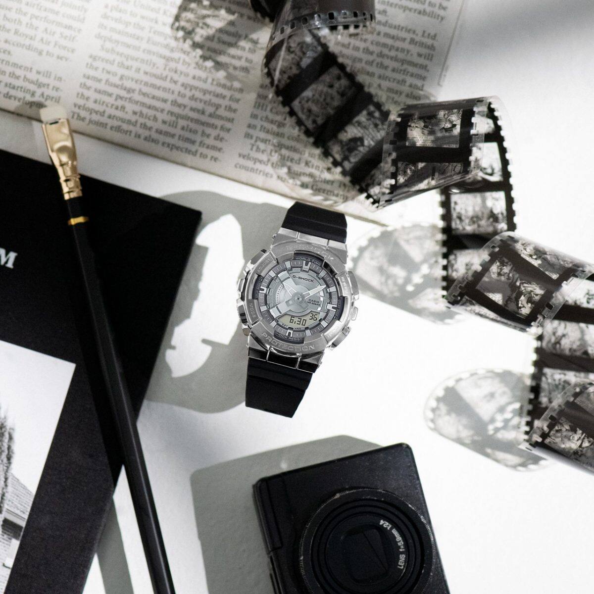 Die GM-S2110-1A Armbanduhr liegt auf einem Tisch neben einem Fotofilm, einer Kamera, einer Zeitung und einem Bleistift.