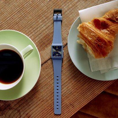 Die flach liegende Uhr MQ-38UC-2A2 auf einem Holztisch neben einem Croissant und einer Kaffeetasse