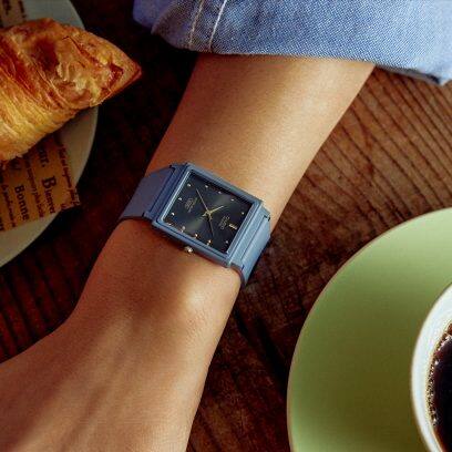 Nahaufnahme des Handgelenks einer Frau, die ihren Arm neben einem Croissant und einer Kaffeetasse auf einem Holztisch abgelegt hat. Am Handgelenk trägt sie die Analoguhr MQ-38UC-2A2.