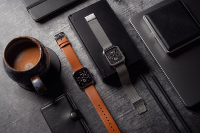 Komplettansicht der flach liegenden CASIO TIMELESS COLLECTION Armbanduhr MTP-M305M-1AV neben dem ähnlichen Modell MTP-M305L-1AV mit orangefarbenem Armband und schwarzem Zifferblatt inmitten verschiedener Schreibtisch- und Fotografie-Utensilien.