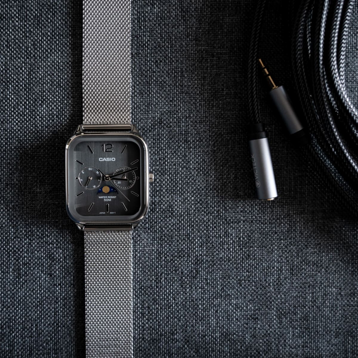 Ansicht der flach liegenden CASIO TIMELESS COLLECTION Armbanduhr MTP-M305M-8AV auf grauem Textiluntergrund neben einem in grauem Textil ummantelten Verlängerungskabel.