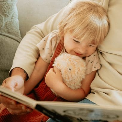 Ein Kind liegt in den Armen eines Erwachsenen, bekommt ein Buch vorgelesen und hält dabei das goldene Moflin im Arm