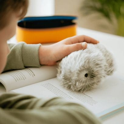 Das Moflin sitzt auf einem aufgeschlagenen Buch und wird von einem Kind gestreichelt