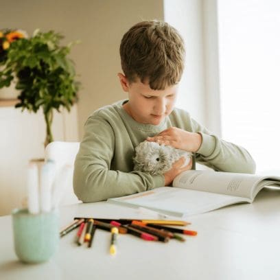 Ein Kind sitzt an einem Tisch auf dem ein aufgeschlagenes Heft und Stifte liegen und streichelt dabei das silberne Moflin