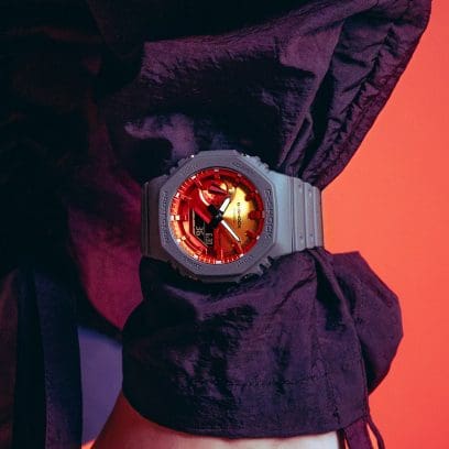 Die GA-2100FL-8A Armbanduhr von G-SHOCK mit orangenem Zifferblatt am Handgelenk. Darunter ist eine violettes Oberteil zu sehen.