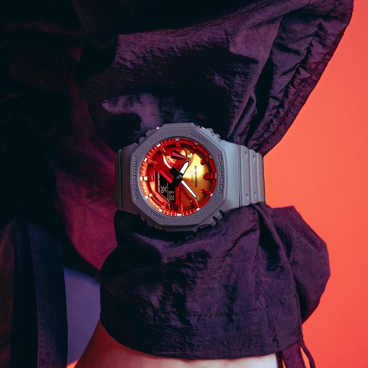 Die GA-2100FL-8A Armbanduhr von G-SHOCK mit orangenem Zifferblatt am Handgelenk. Darunter ist eine violettes Oberteil zu sehen.