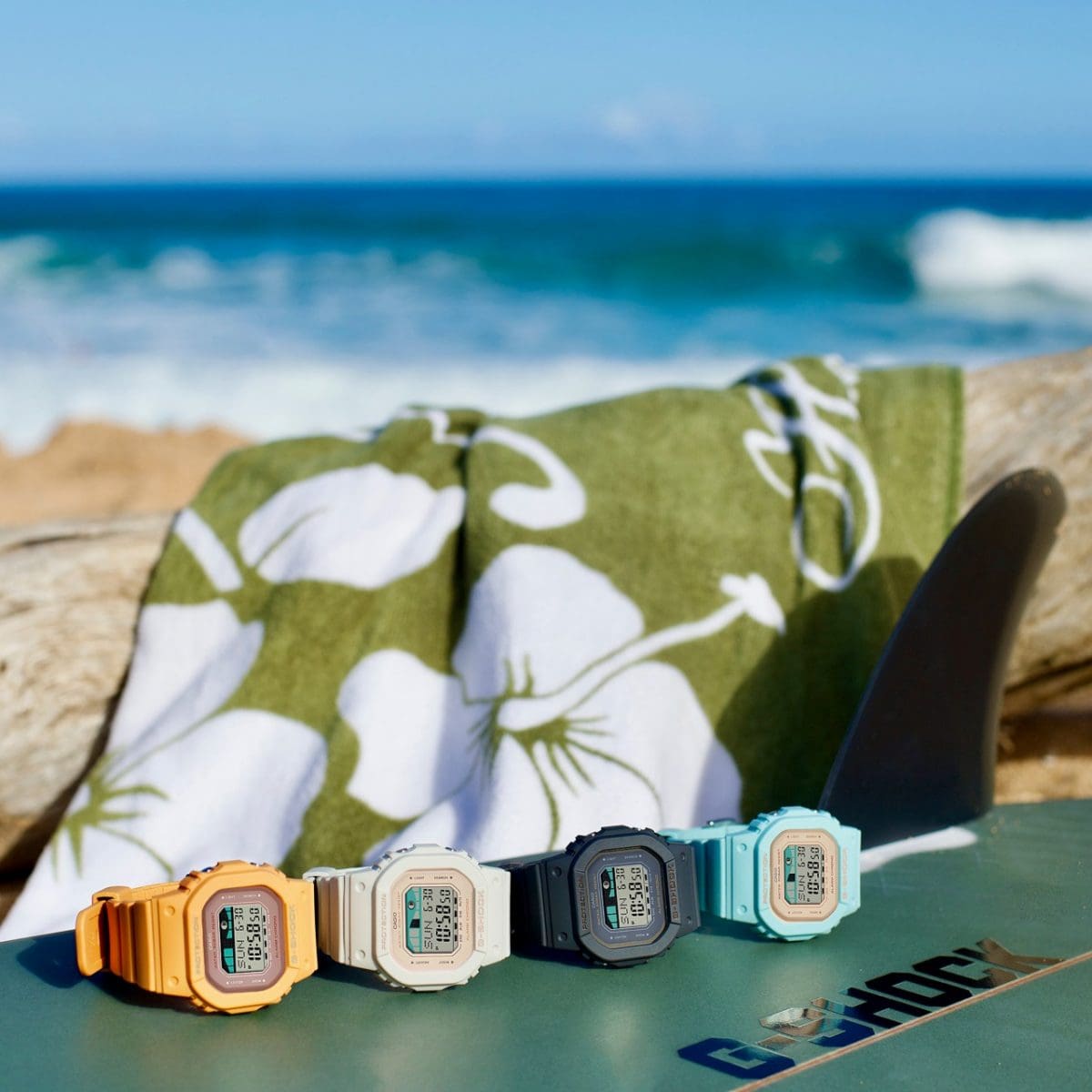Vier Surfer-Digitaluhren von G-SHOCK: die orangefarbene GLX-S5600-4, die GLX-S5600-7B mit beige-weißem Armband, die GLX-S5600-1 mit grauem Armband und die GLX-S5600-3 in türkis auf einem Surfboard am Strand