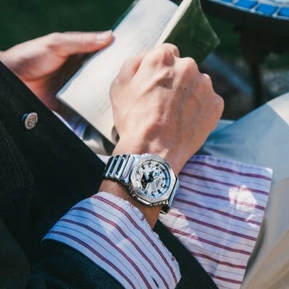 Nahaufnahme der G-SHOCK GM-2100YM-8A Armbanduhr am Handgelenk eines Mannes, der ein Buch in den Händen hält.