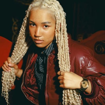 Eine junge Frau  mit langen hellblonden geflochtenen Haaren trägt die GM-2100YMG-9A9ER am linken Handgelenk in Kombination mit einer bordeauxroten Jacke, einem schwarz-weiß gestreiften Hemd und zwei goldenen Ringen an ihren Fingern. Sie sitzt dabei auf einem roten Sessel in einem dunklen Kinosaal.