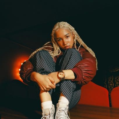 Eine junge Frau  mit langen hellblonden geflochtenen Haaren sitzt in angewinkelter Haltung auf einem hölzernen Boden. Sie trägt dabei die GM-2100YMG-9A9ER am linken Handgelenk. Sie kombiniert die Uhr mit  einer bordeauxroten Jacke und einer gestreiften Hose. 