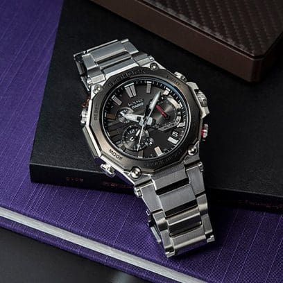 Die CASIO MTG-B2000D-1A Armbanduhr mit schwarzem Zifferblatt, silbernem Edelstahlgehäuse und silbernem Edelstahlarmband liegt auf mehreren Notizbüchern in lila und schwarz