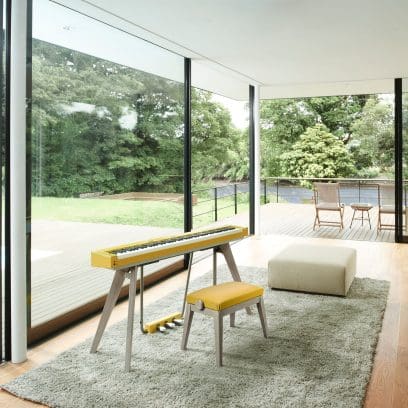 Fotografía del piano digital CASIO Privia PX-S7000 con banqueta, en el color Harmonious Mustard, en un salón de estilo moderno con ventanales de suelo a techo y vistas a un jardín con terraza.