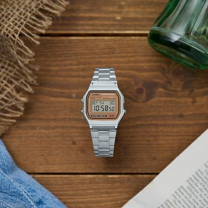 Reloj digital A158WEA-9 con correa plateada y marco dorado sobre mesa de madera rodeada de diversos objetos