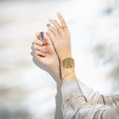 Dos manos femeninas elegantemente cruzadas: la mano derecha sostiene un pequeño frasco de perfume, mientras que el reloj de pulsera A168XESG-9A se acomoda en la muñeca izquierda.