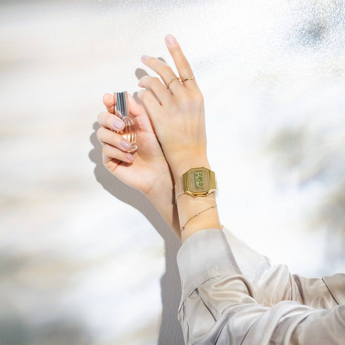 Dos manos femeninas elegantemente cruzadas: la mano derecha sostiene un pequeño frasco de perfume, mientras que el reloj de pulsera A168XESG-9A se acomoda en la muñeca izquierda.