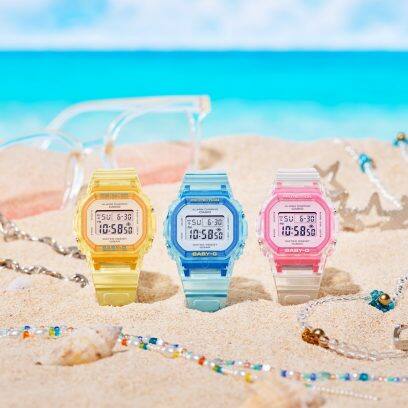 Los relojes de pulsera digitales amarillo, azul claro y rosa de Baby-G en la arena junto a collares de colores y gafas de sol brillantes