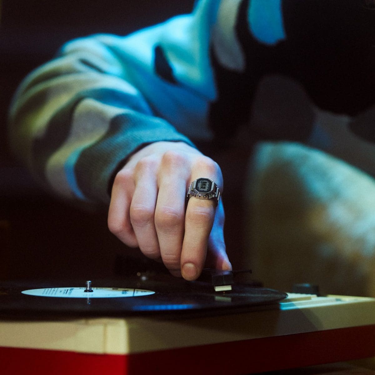 Primer plano de una mano moviendo la aguja de un tocadiscos. La persona lleva el CASIO Ringwatch CRW-001 con pantalla digital en el dedo índice.