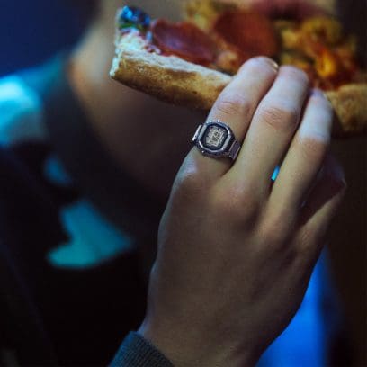 Primer plano de una mano sosteniendo una pizza. El CASIO CRW-001 Ringwatch está en el dedo índice.