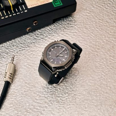 El reloj de pulsera GMA-S2110P con caja metálica negra y correa de resina gris y negra sobre una alfombra beige. 