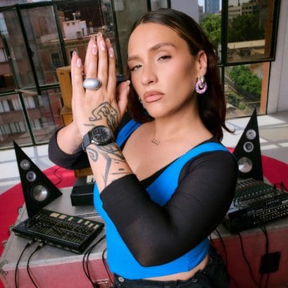 Lia Kali, con un top azul y negro, mira a la cámara y junta las manos delante de la cara. En la muñeca lleva el reloj de pulsera analógico negro y gris GM-S2110B-8A de G-SHOCK.