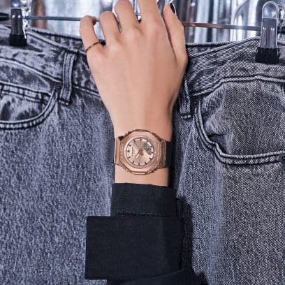 Primer plano del reloj analógico color rosa GM-S2110PG-1A4 en la muñeca de una mujer que sostiene un par de jeans en una percha.