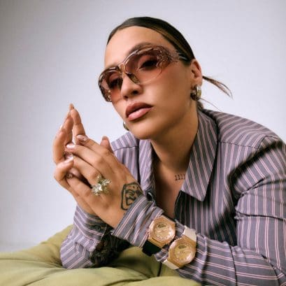 Lia Kali se apoya en el codo y mira directamente a la cámara, lleva gafas de sol de color rosa y dos relojes analógicos G-SHOCK en la muñeca con biseles metálicos de oro rosa.