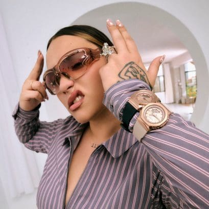 La rapera Lia Kali, con una blusa a rayas y gafas de sol de color rosa, guiña el ojo a la cámara y se lleva las manos a la cabeza. Lleva dos relojes de pulsera GM-S2110 con correas negras y beige en la muñeca.