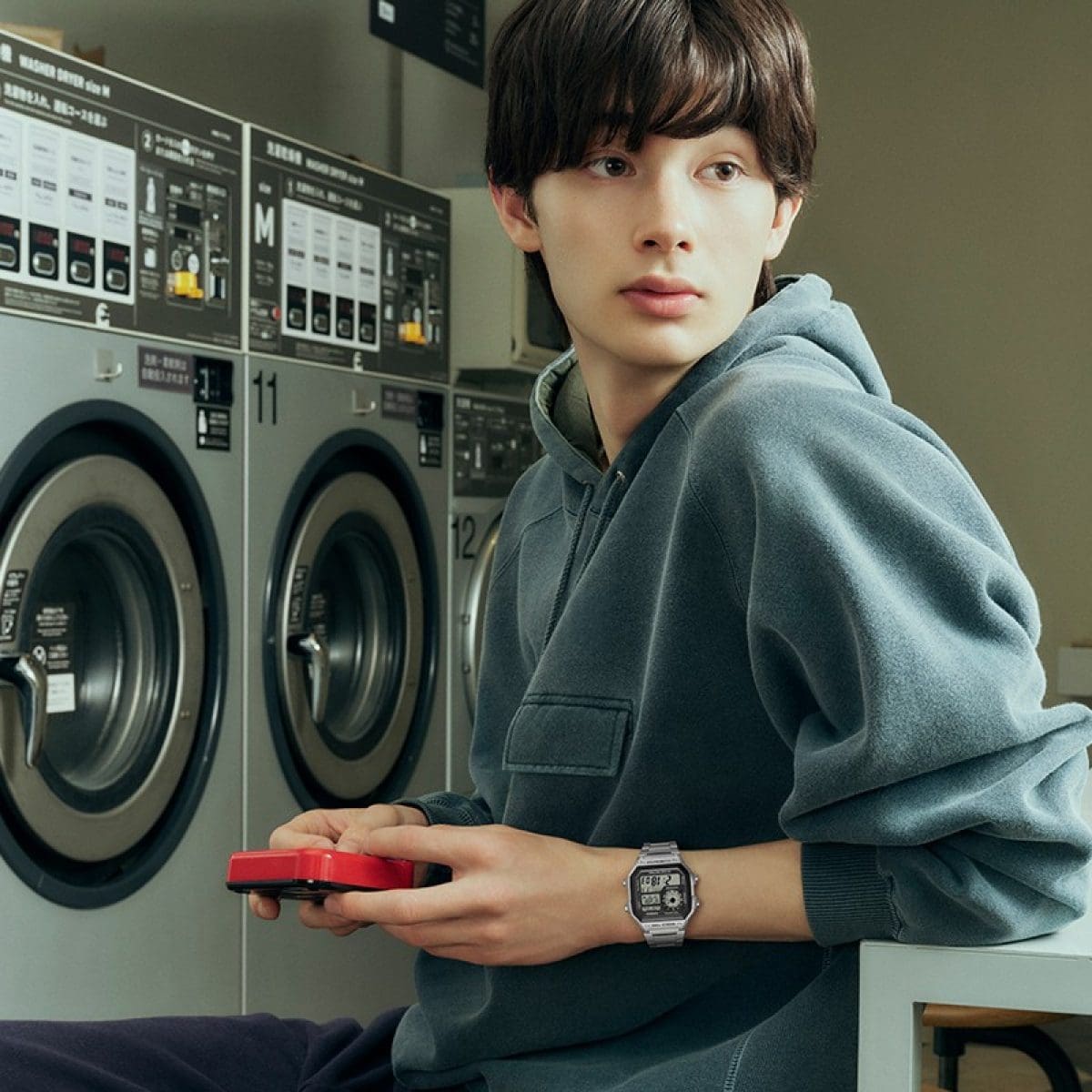 A boy with brown hair in a laundromat wearing a blue-grey hoodie and the CASIO Timeless Collection watch AE-1200WHD-1AV and holding his phone with a red case in his hands.