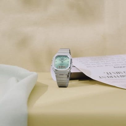 The CASIO VINTAGE COLLECTION AQ-240E-2AEF wristwatch is displayed against a cream-colored background with a folded newspaper in the background.