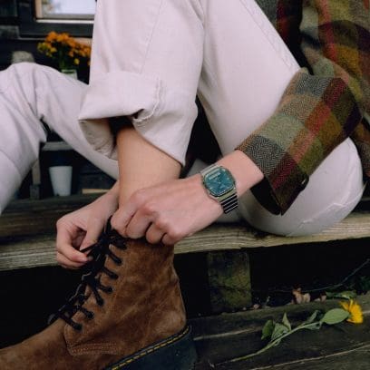 Eine Frau trägt die AQ-240E-3AEF am Handgelenk und schnürt sich gerade die Schuhe zu. Sie trägt eine beige Hose und eine karierte Wolljacke