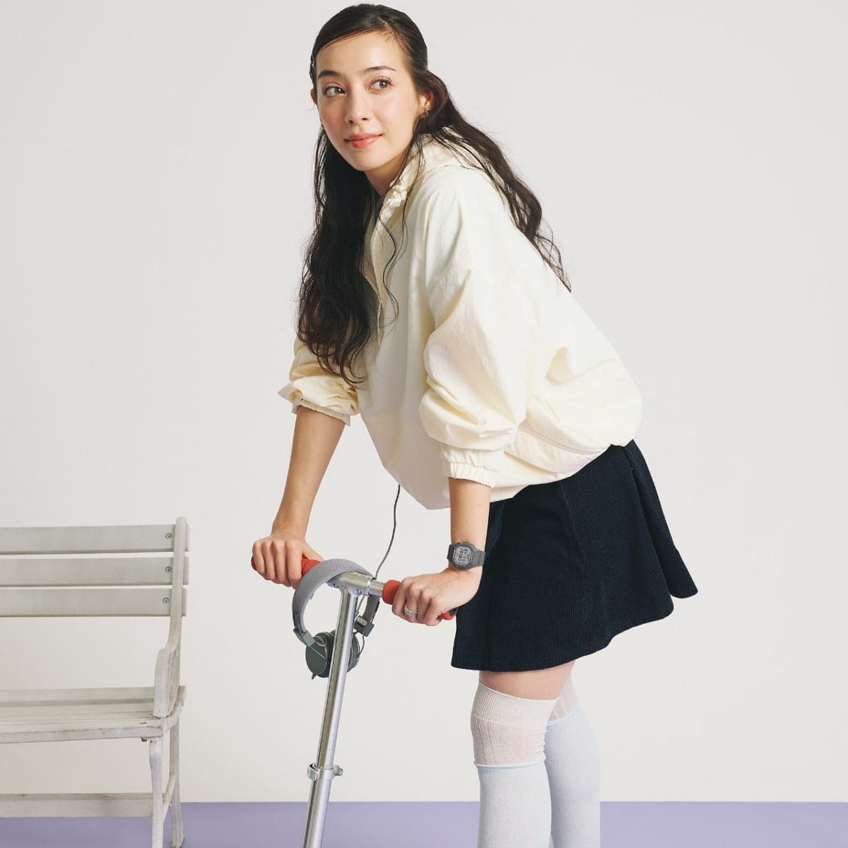 A woman wearing a dark skirt and a light sweater is standing on a scooter and wearing the BABY-G digital watch BGD-565SC-1ER on her wrist.