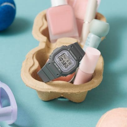 The grey BABY-G wristwatch BGD-565SC-1ER lies in a cardboard cup holder, along with other cosmetic items.