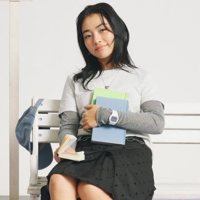 A woman sits on a bench holding pastel-colored books, while wearing a white BGD-565SC-2BER with a blue dial on her wrist.